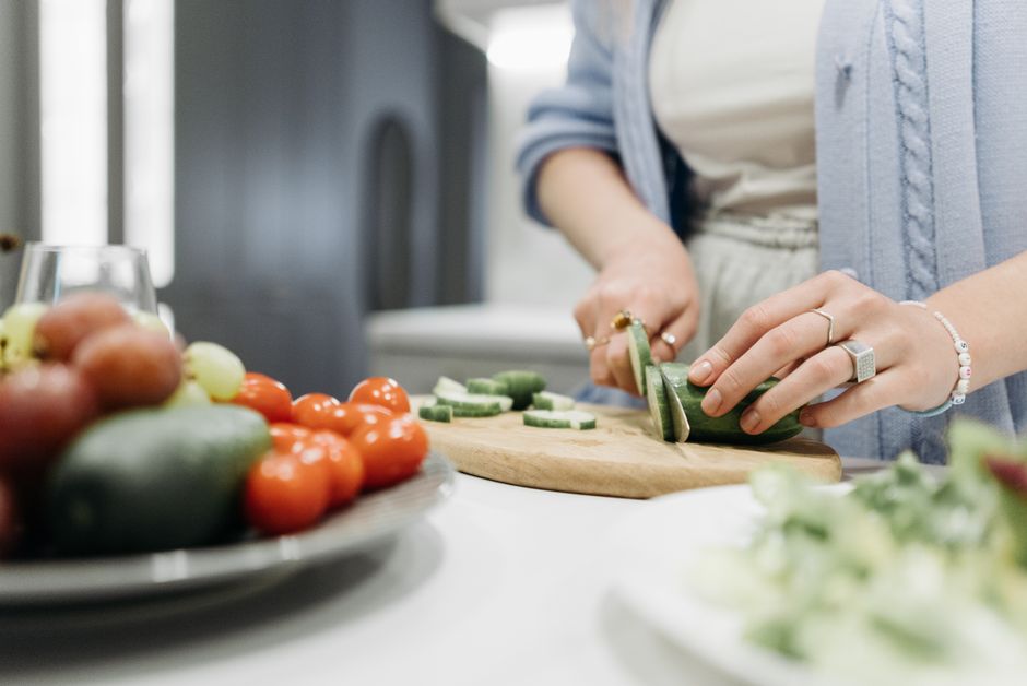 Femme coupant un concombre sur une planche, avec des légumes frais en arrière-plan, illustrant la cuisine sans bagues.