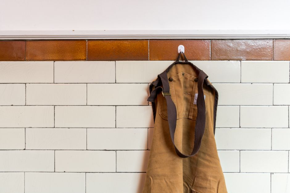 Tablier en toile suspendu à un crochet sur un mur carrelé blanc et marron, créant un coin pratique pour objets personnels.