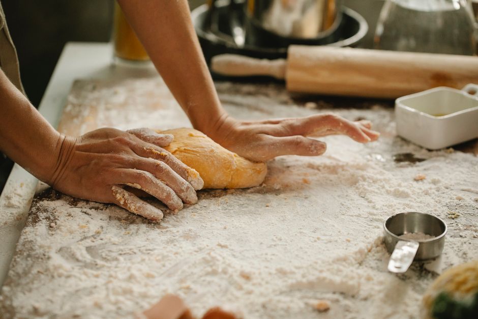 Mains pétrissant une pâte sur un plan de travail couvert de farine, avec des ustensiles de cuisine en arrière-plan.