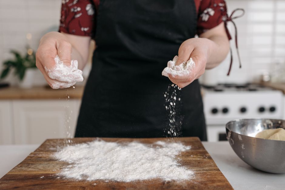 Femme en tablier saupoudrant de la farine sur un plan de travail en bois avant de cuisiner.