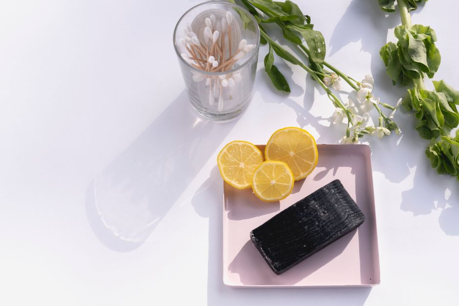 Barre de savon noir sur un plateau rose, tranches de citron et vase avec des bâtonnets en bois et des fleurs blanches.