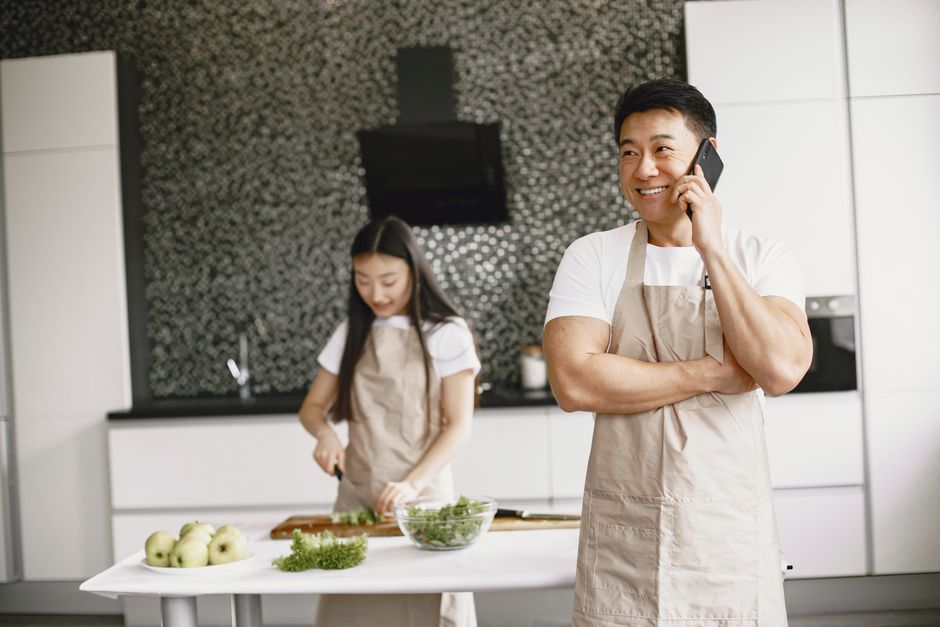 Homme au téléphone souriant dans une cuisine, tandis qu'une femme coupe des légumes en arrière-plan.