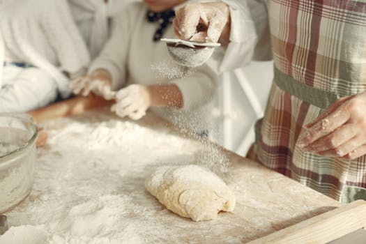 Un adulte saupoudre de la farine sur une pâte à biscuits, avec des enfants en arrière-plan, dans une cuisine.